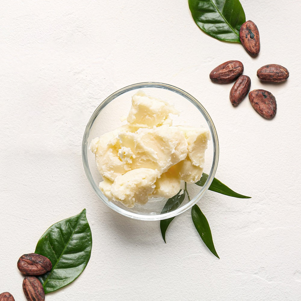 Top View Of Cocoa Butter In Glass Bowl Sorrounded By Eight Cocoa Beans And Leaves | Bulk Cosmetic Butters | Brightpack Raw Materials